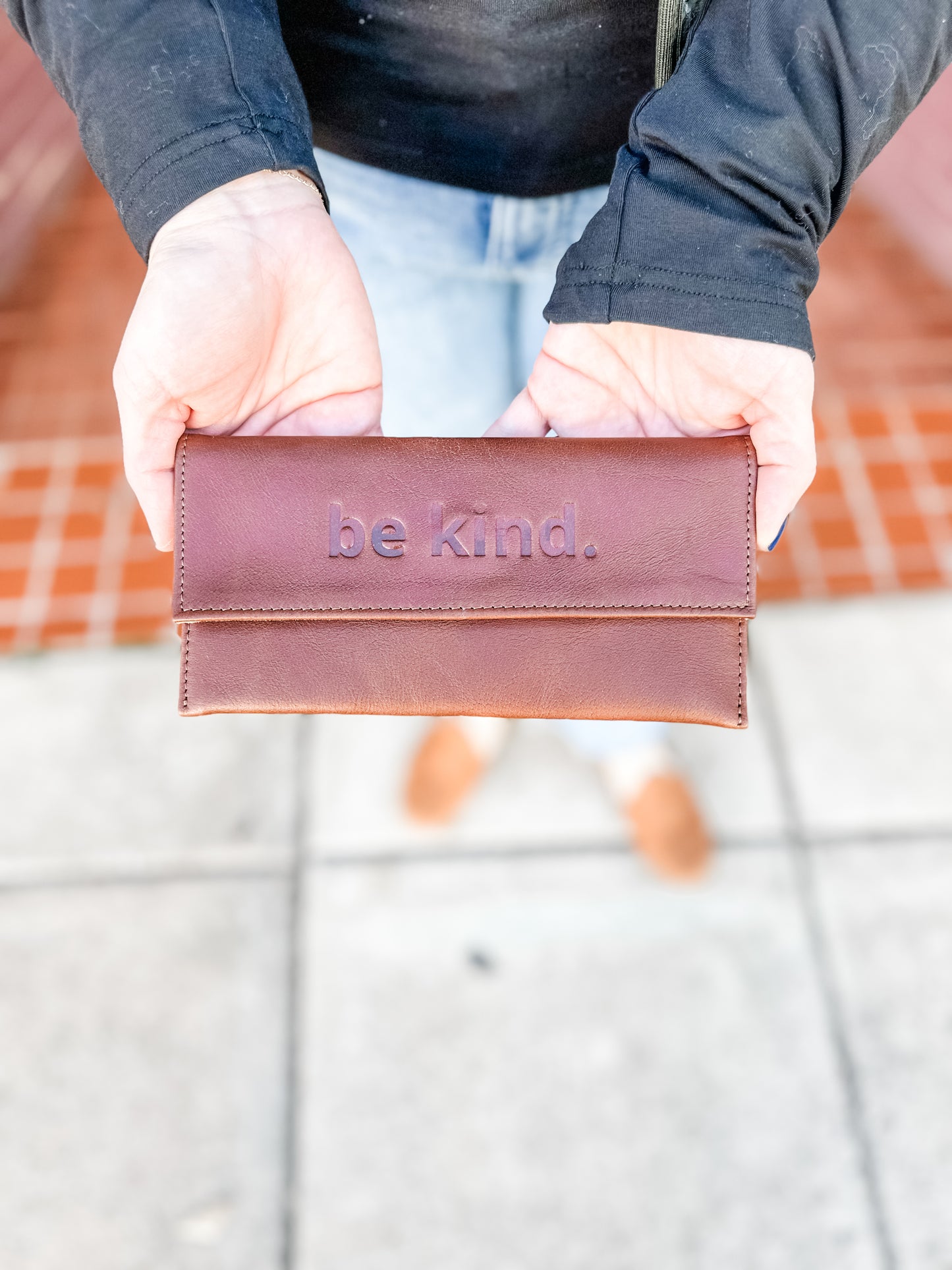 A person holding a brown leather sunglasses case with the text 'be kind' embossed on the front.