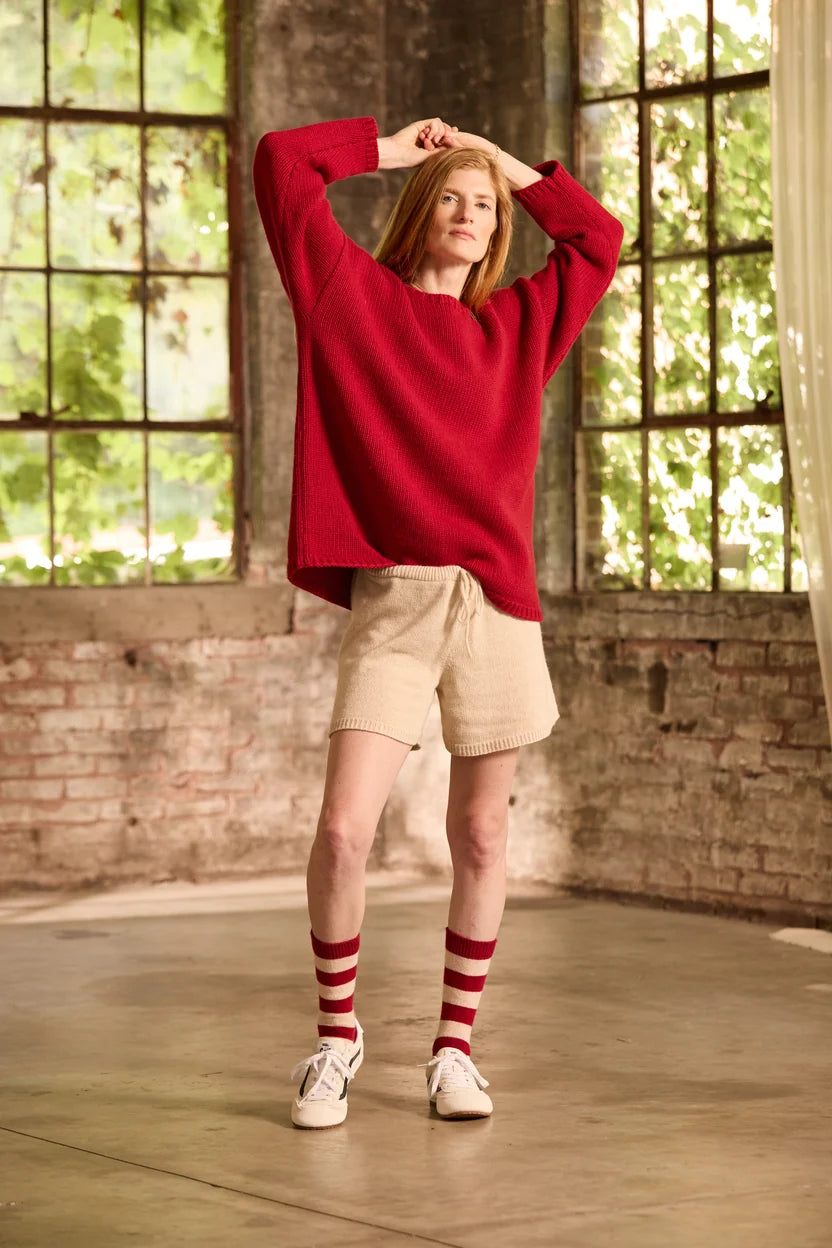 A woman stands in a rustic indoor loft with large windows and brick walls, wearing a loose red knit sweater, beige shorts, red-and-white striped socks, and white sneakers.